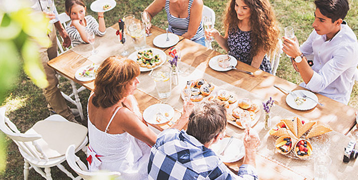 Eine große Familie sitzt im sommerlichen Garten an einem gedeckten Tisch und isst. 