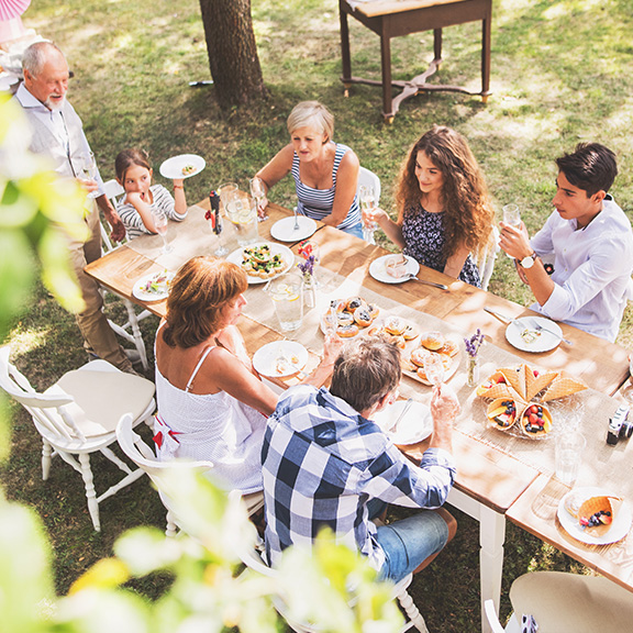 Eine große Familie sitzt im sommerlichen Garten an einem gedeckten Tisch und isst. 
