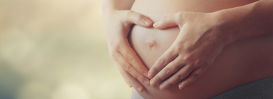 Eine Schwangere formt mit den Händen ein Herz auf ihrem Bauch.