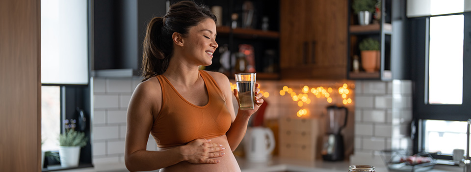 Eine Schwangere steht mit einem Glas Wasser in der Küche. Ihre rechte Hand liegt auf ihrem Bauch.
